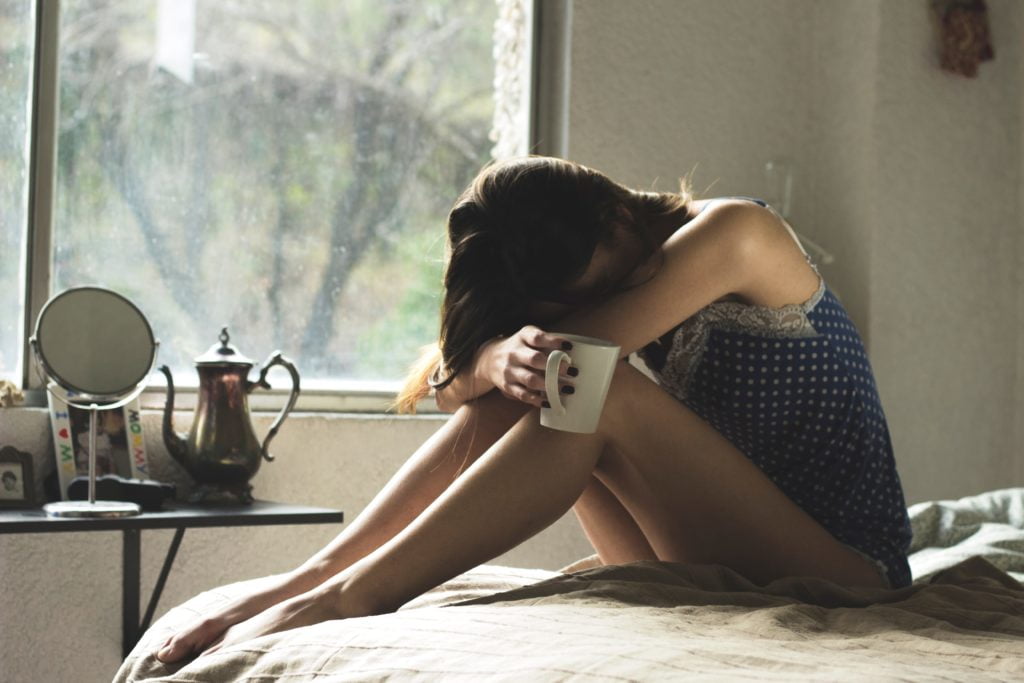 Image of a woman in front of a window on a bed holding a coffee cup who looks sick